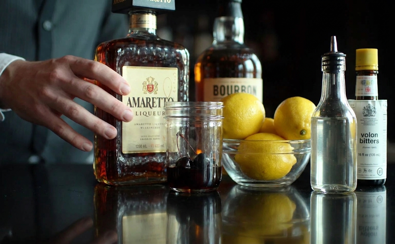 Amaretto, bourbon, fresh lemons, simple syrup, bitters, and a jar of cocktail cherries arranged on a clean kitchen counter