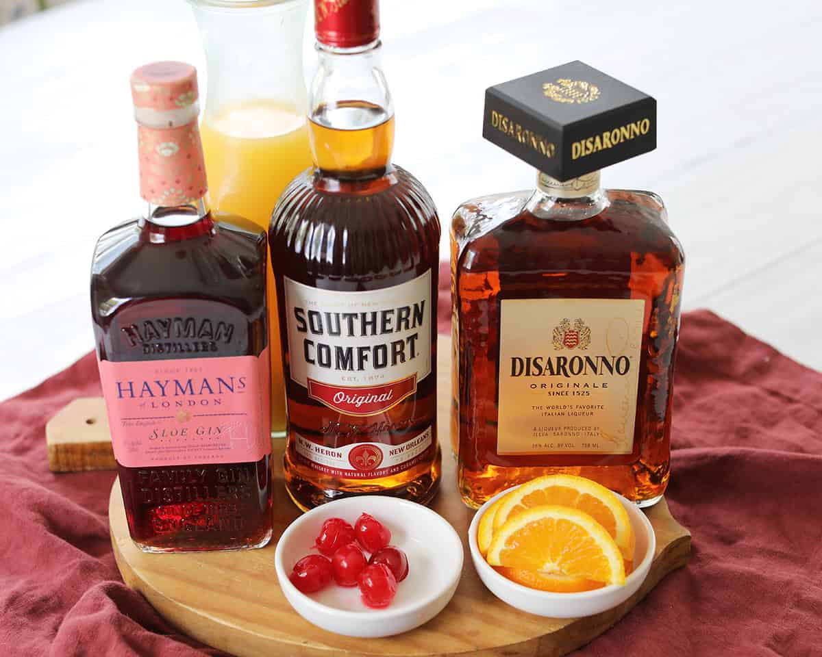 Amaretto, sloe gin, Southern Comfort, fresh oranges, a jigger, and a cocktail shaker arranged on a clean kitchen counter with warm natural light