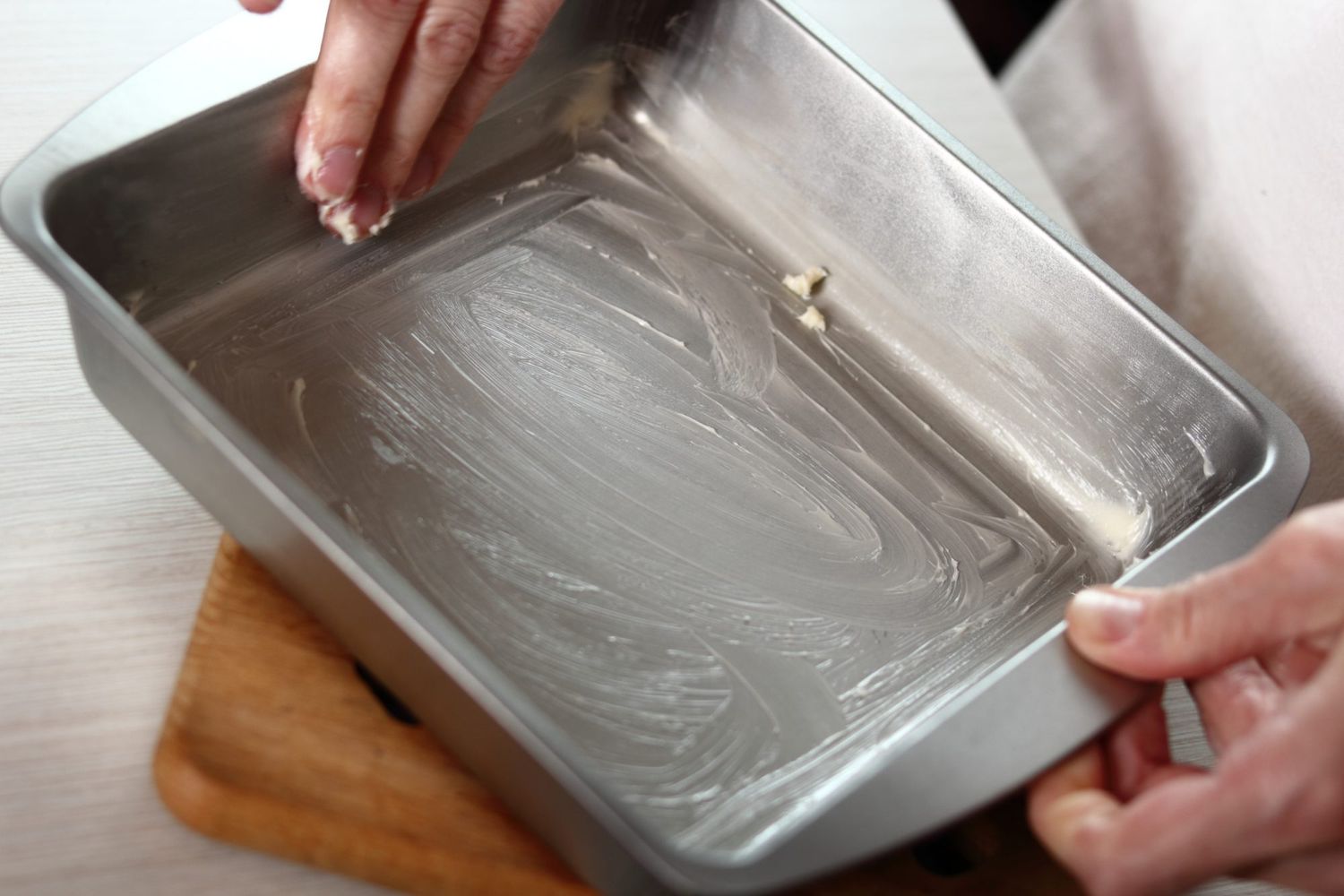 An 8x8-inch baking dish being buttered on a light stone counter with a stick of butter and pastry brush nearby