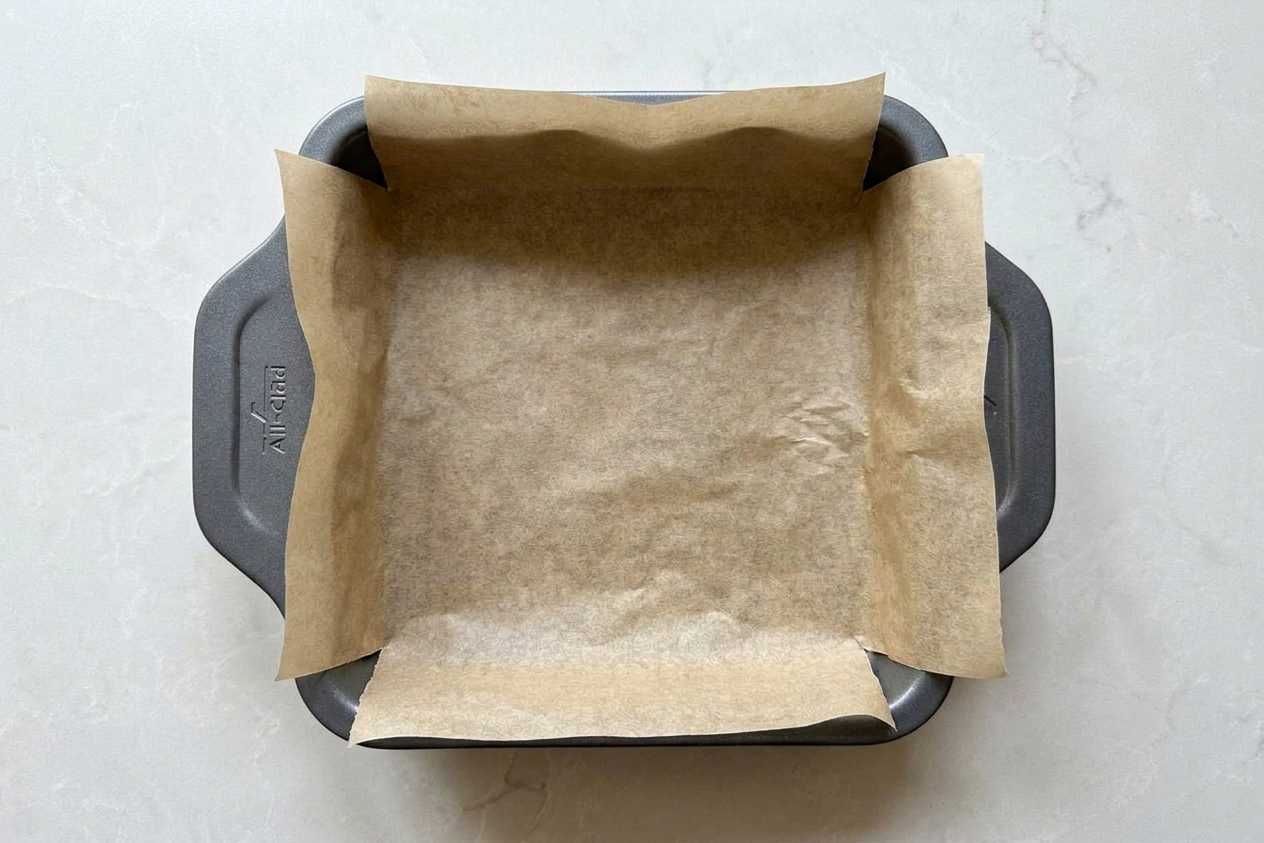 An 8x8-inch metal baking pan lined with parchment paper with overhang on a kitchen counter