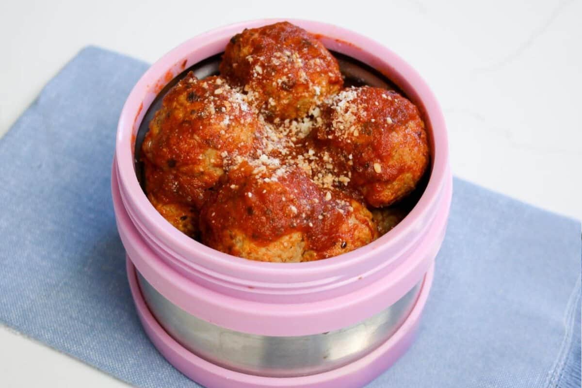 An airtight glass container filled with grape jelly meatballs and sauce cooling on a kitchen counter with the lid set beside it