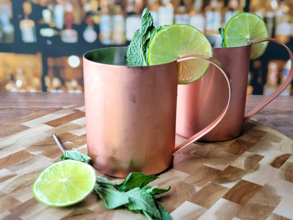 An icy copper mug filled with a pale amber whiskey and ginger cocktail, crushed ice piled high, a lime wheel on the rim, and condensation beading on the mug on a kitchen counter