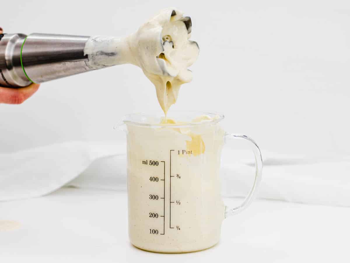 An immersion blender blending a thickening vegan mayonnaise in a tall jar on a clean kitchen counter