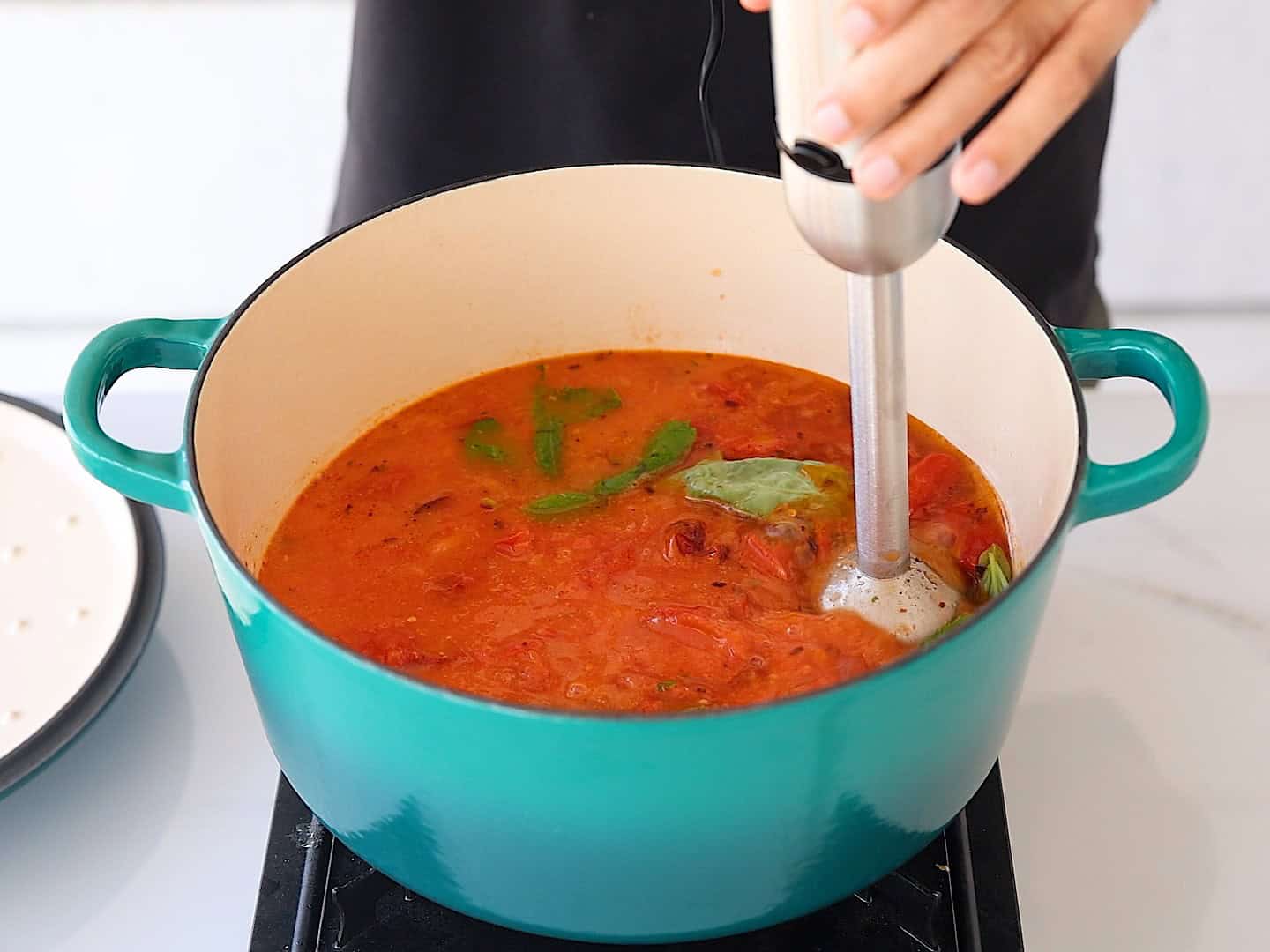 An immersion blender blending hot tomato soup in a large pot on the stove, soup turning smooth and creamy