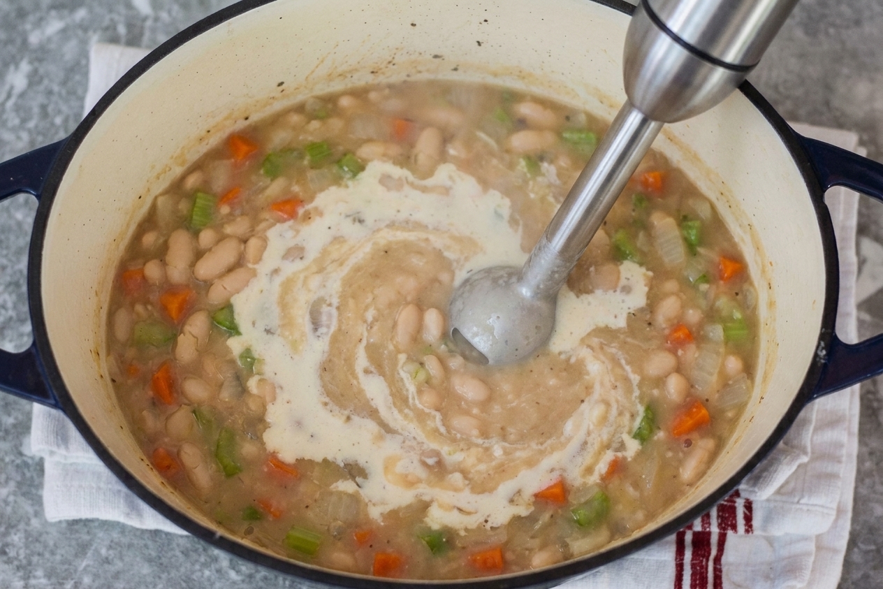 An immersion blender partially submerged in a pot of cannellini bean soup, creating a creamy swirl