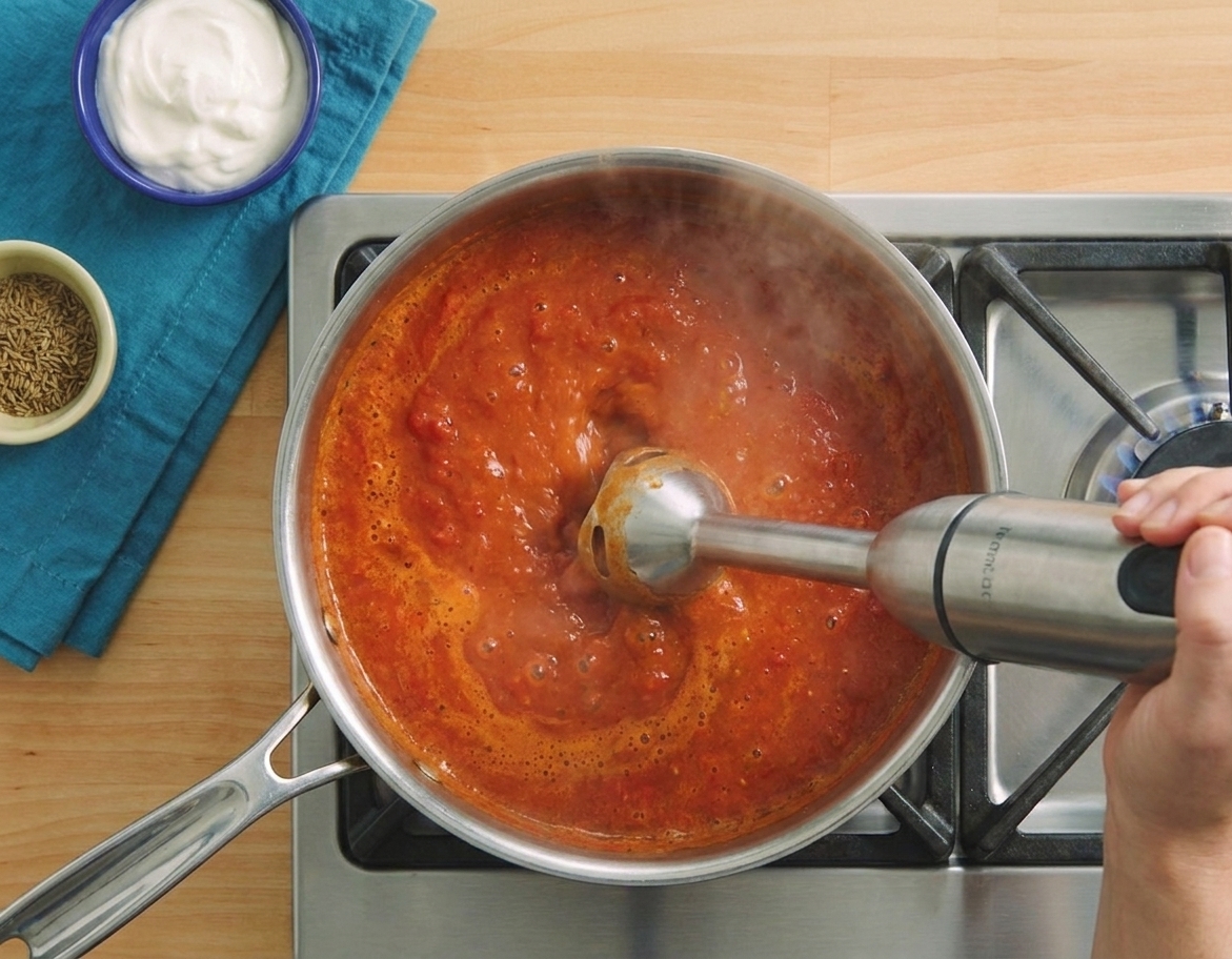 An immersion blender pureeing a simmering tomato spice sauce in a pot on the stove