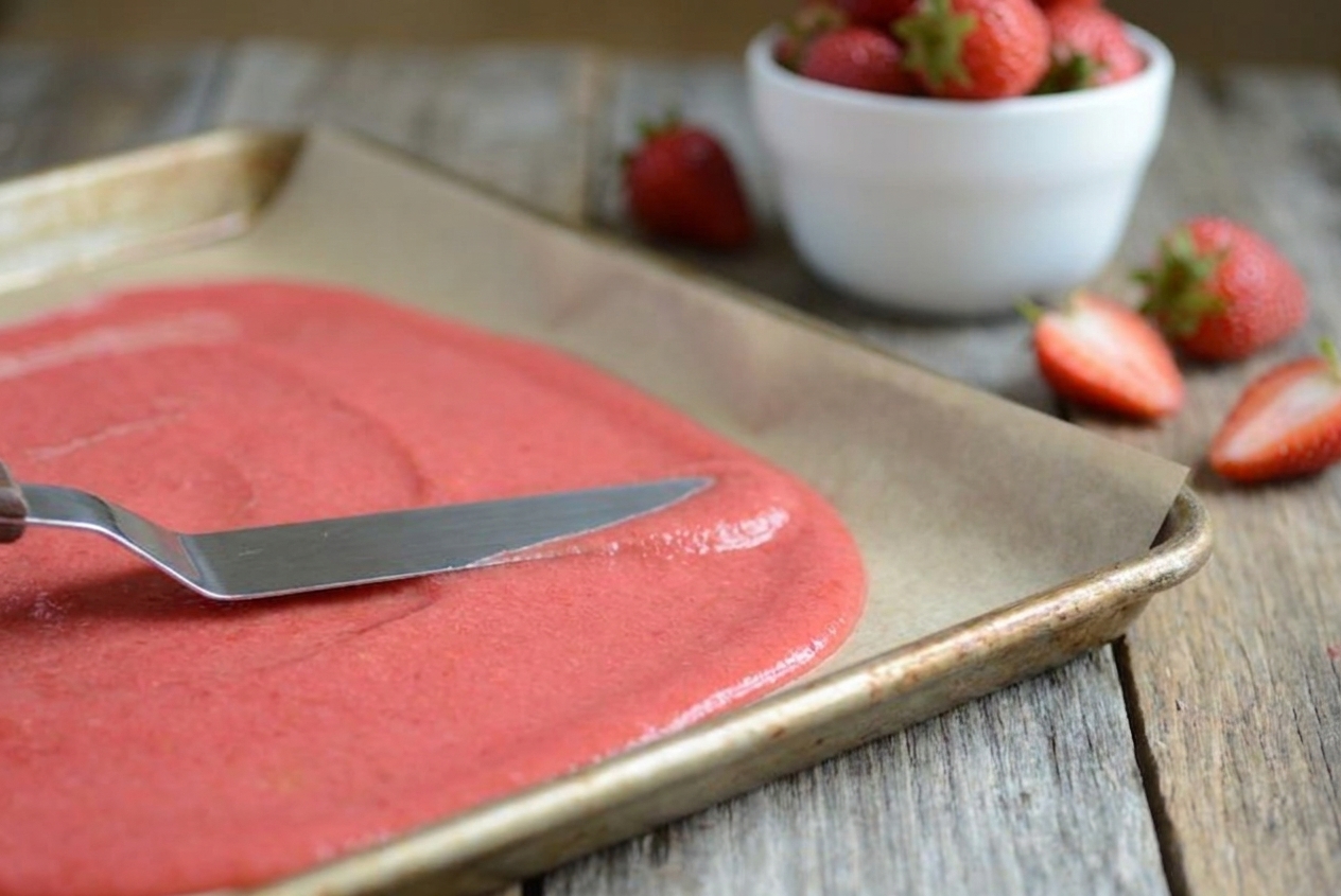 An offset spatula spreading pink fruit puree into an even layer on a parchment-lined sheet pan