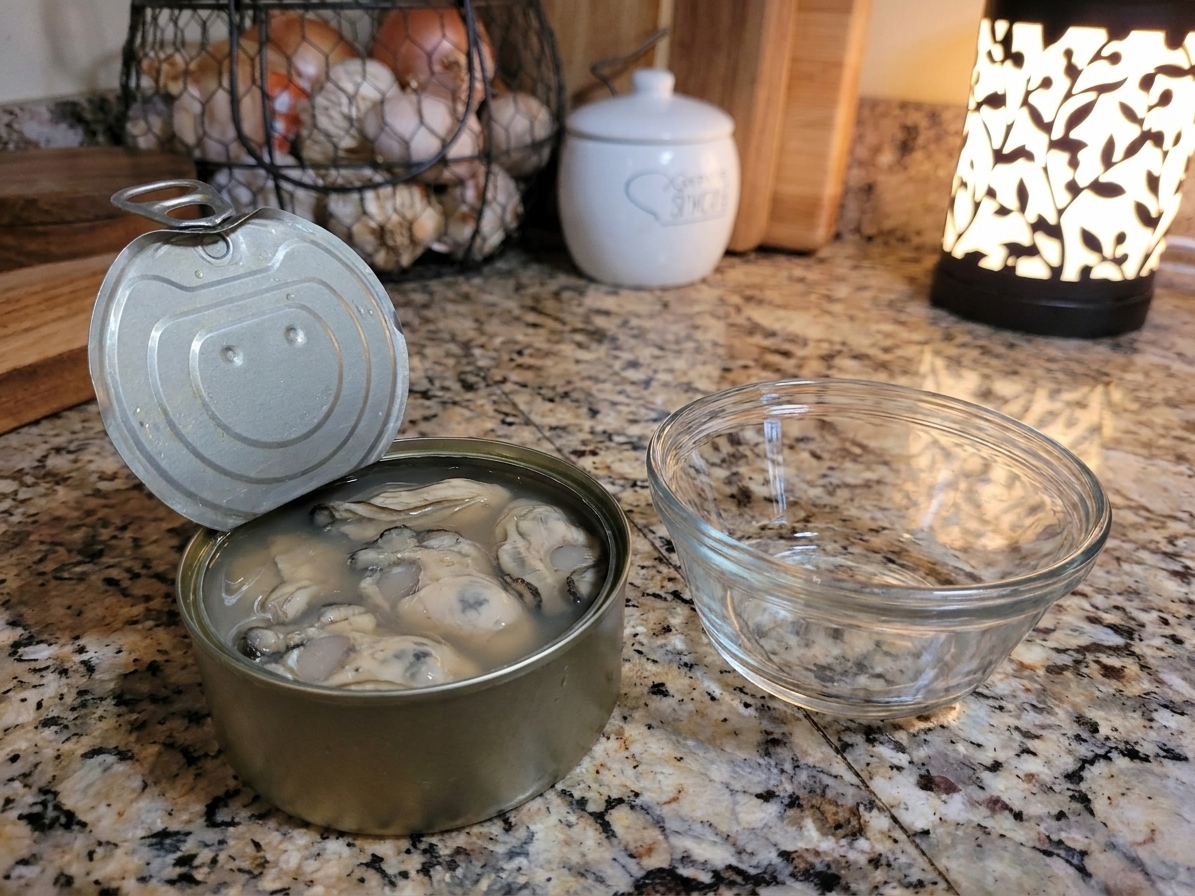 An opened can of oysters with the oyster liquor visible, set on a kitchen counter next to a small bowl