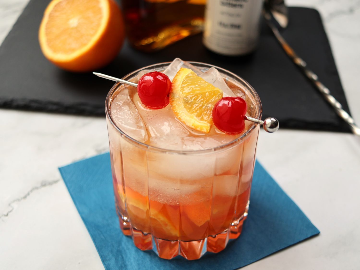 An orange slice and a maraschino cherry on a small plate beside a cutting board on a kitchen counter
