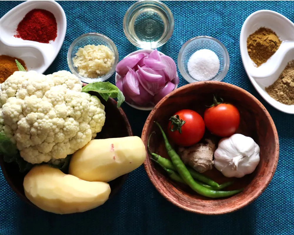 An overhead photo of ingredients for aloo gobi arranged on a kitchen counter, including cauliflower, potatoes, onion, tomatoes, ginger, garlic, spices in small bowls, and a lemon
