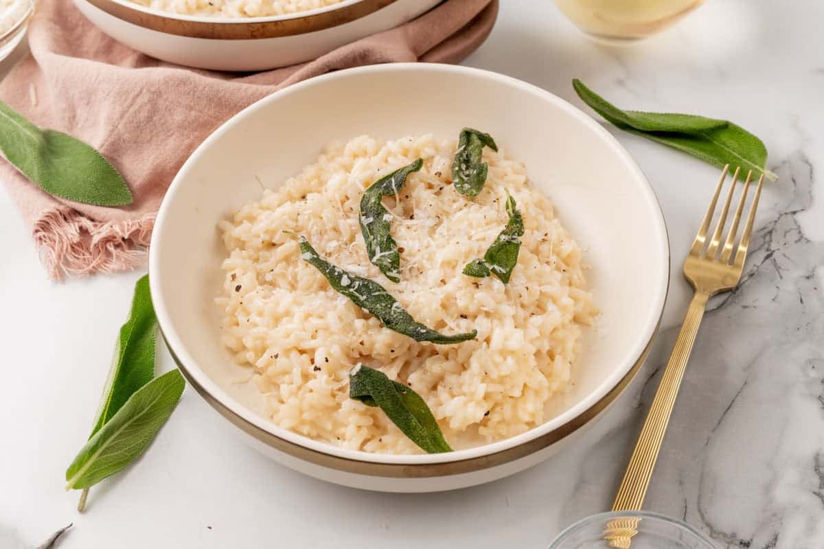 Arborio rice in a small bowl beside grated Parmesan and fresh sage on a wooden cutting board