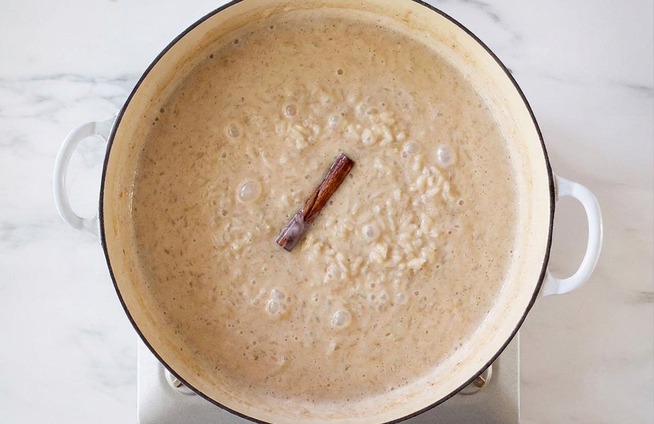 Arroz con leche gently simmering in a saucepan on a stovetop with small bubbles and a cinnamon stick floating on top