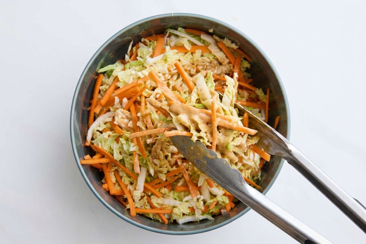 Asian coleslaw being tossed with metal tongs as the sesame-ginger dressing coats the cabbage and carrots