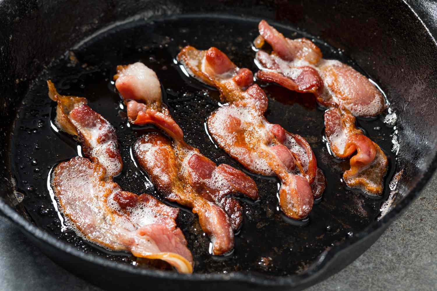 Bacon pieces sizzling in a cast iron skillet with rendered fat pooling and edges turning golden