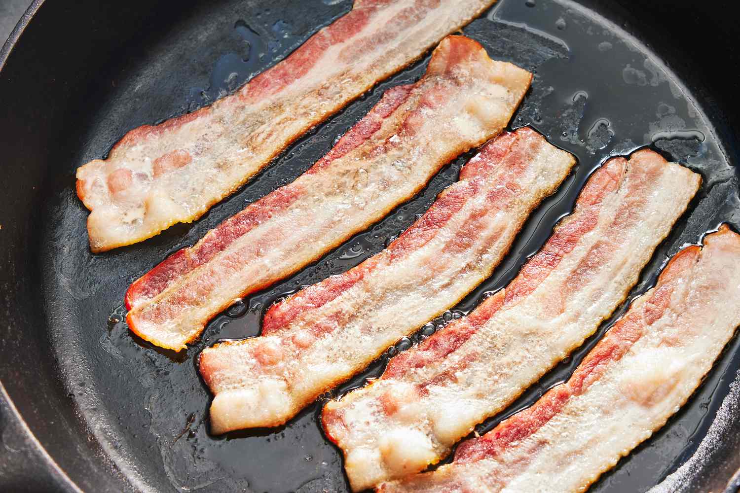 Bacon strips sizzling and turning crisp in a black skillet on a stovetop under warm kitchen lighting