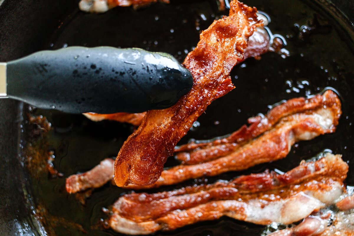 Bacon strips sizzling in a black skillet on a home stove with a wooden spoon nearby