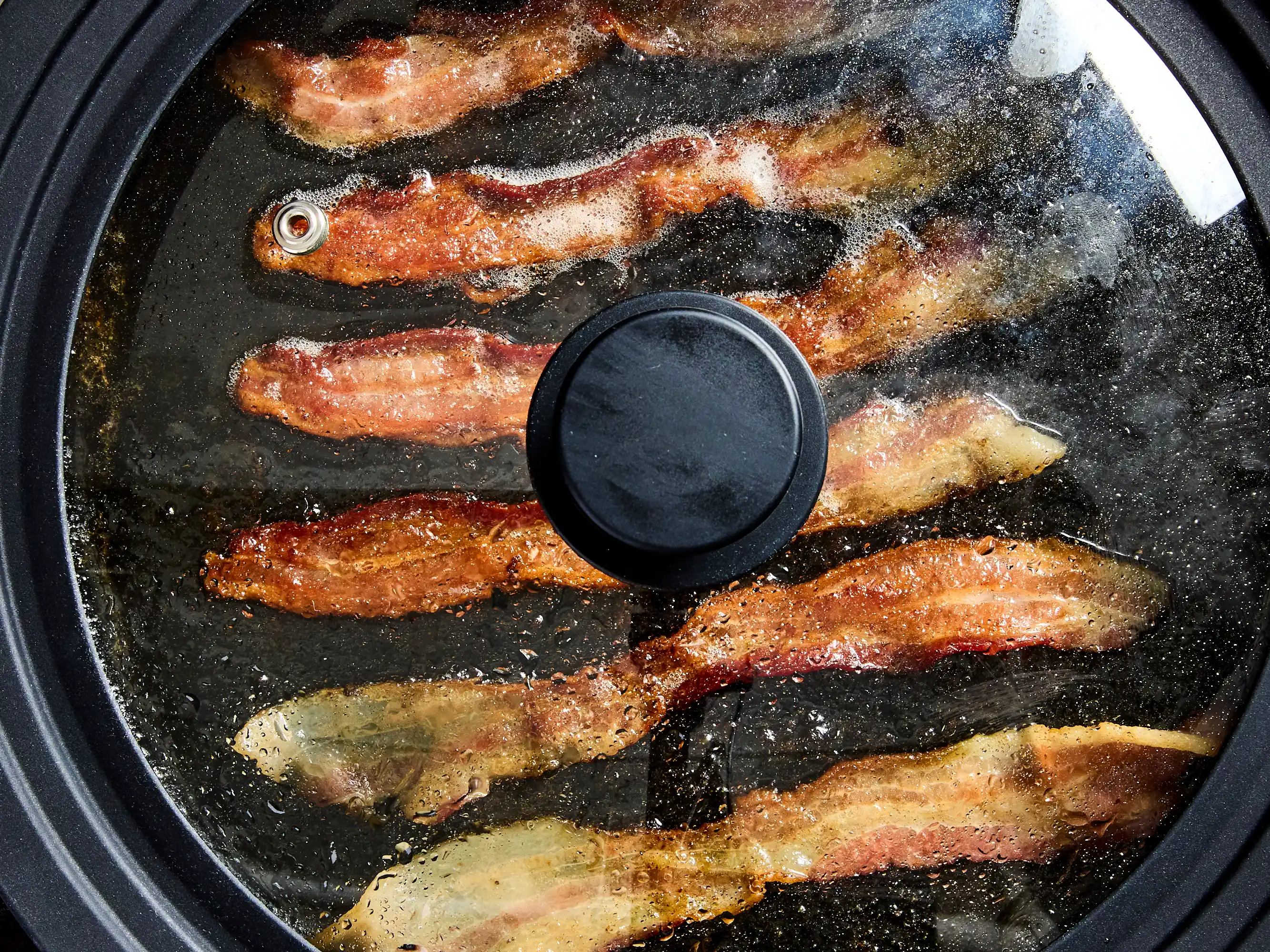 Bacon strips sizzling in a stainless steel skillet on a stovetop with crisp edges forming