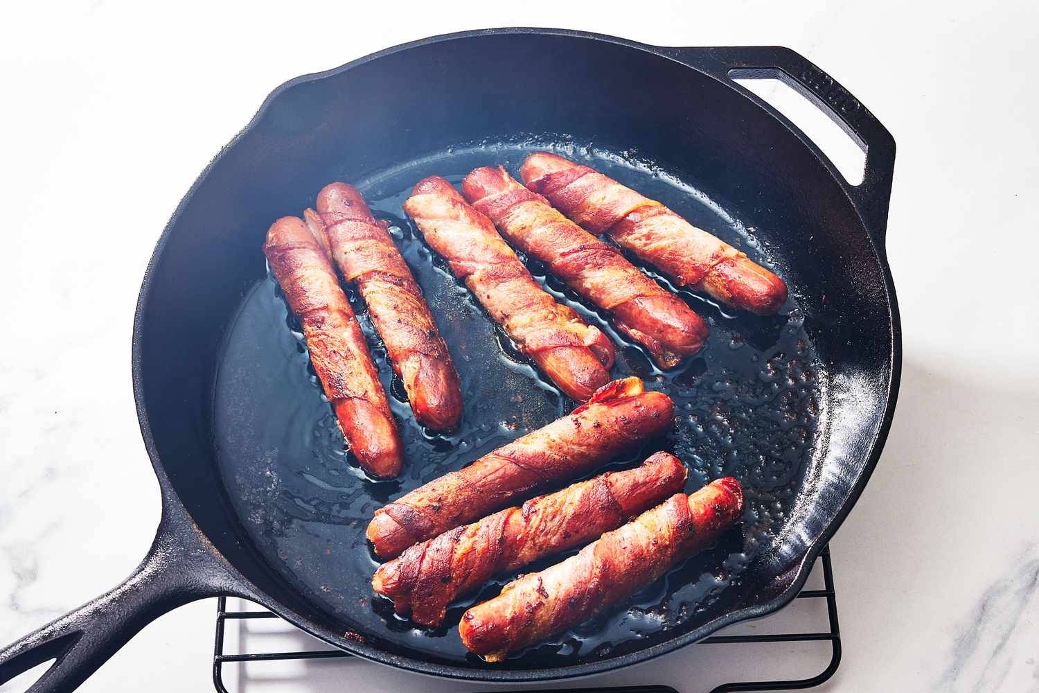 Bacon-wrapped hot dogs sizzling in a cast iron skillet on a stovetop, with tongs turning them as the bacon crisps