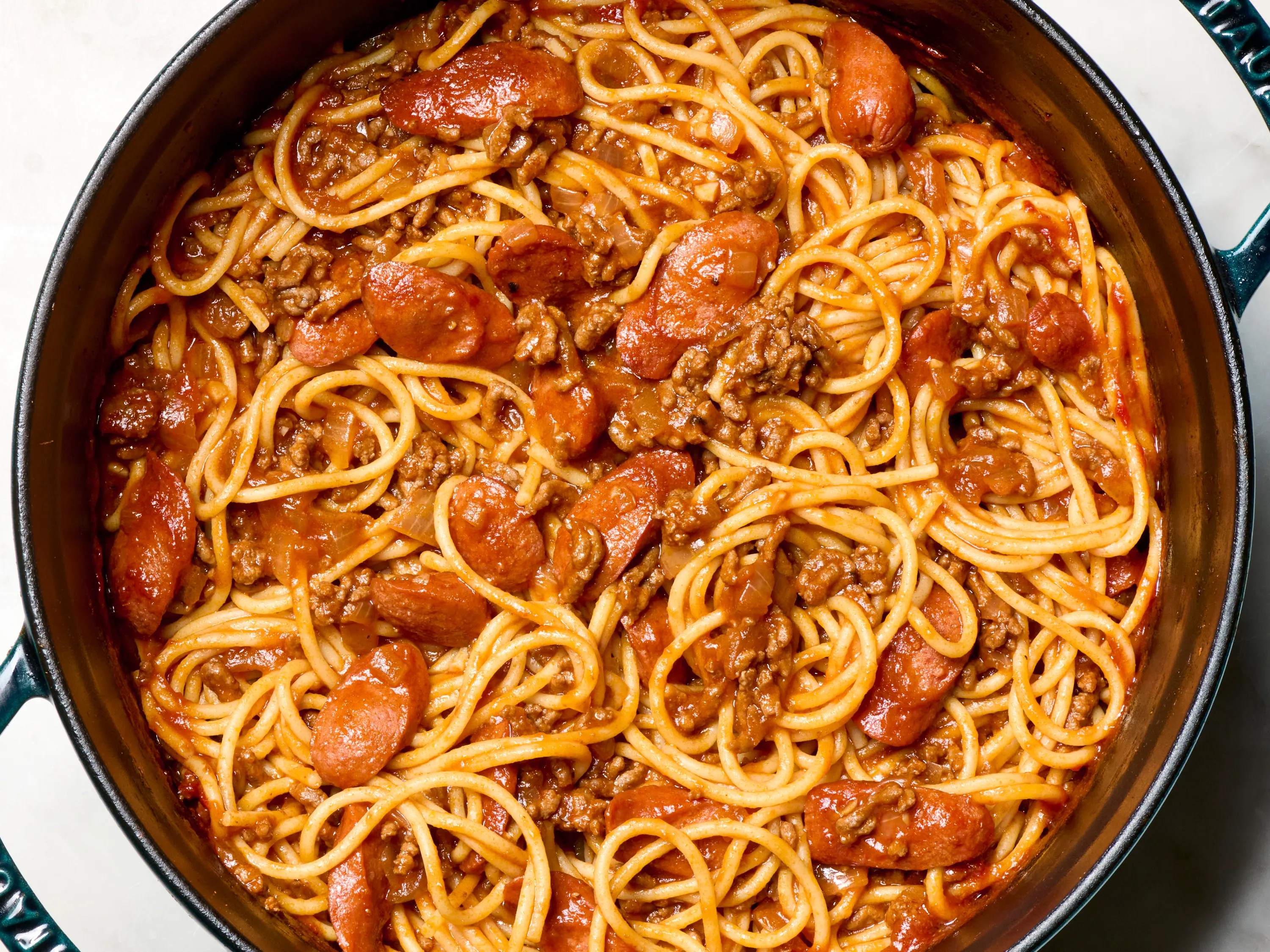 Banana ketchup, tomato sauce, ground meat, hot dogs, onions, garlic, spaghetti, and shredded cheddar arranged on a kitchen counter with warm natural light