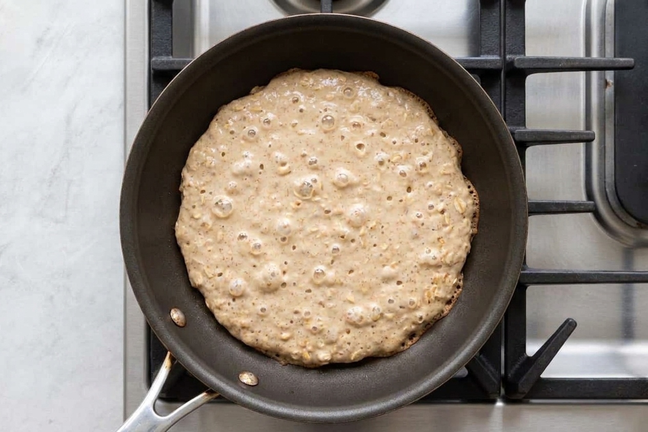 Banana oat pancake batter cooking in a nonstick skillet with bubbles forming on the surface, viewed from above