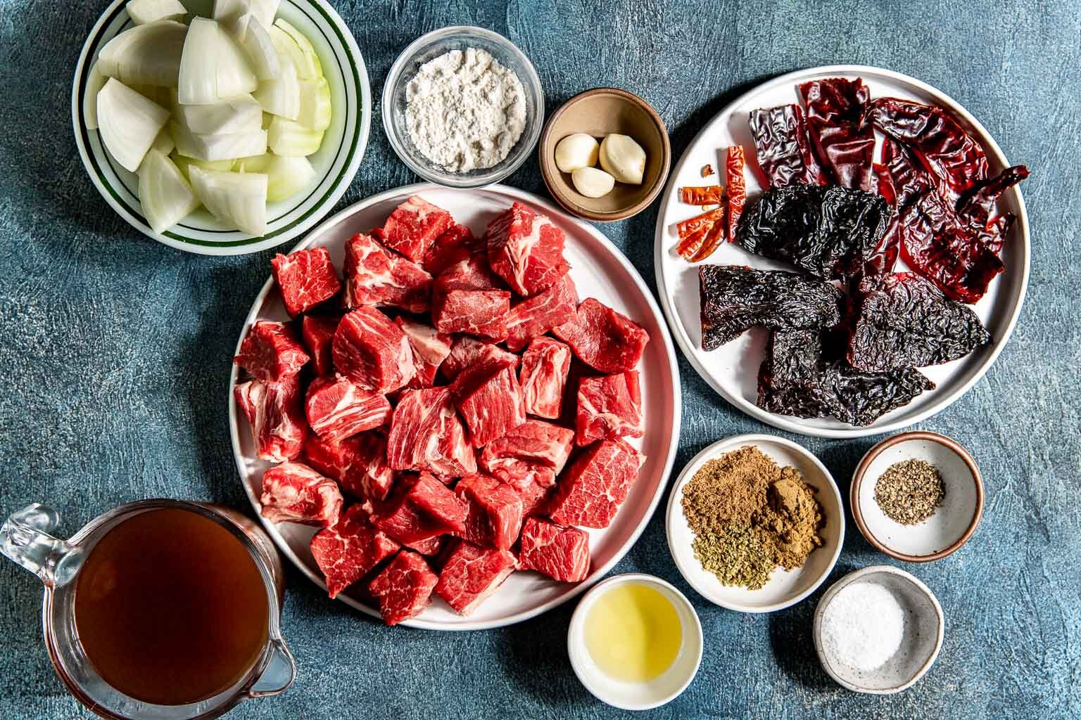 Beef chuck cubes, dried guajillo and ancho chiles, onion, garlic, and small bowls of spices arranged on a kitchen counter ready for cooking