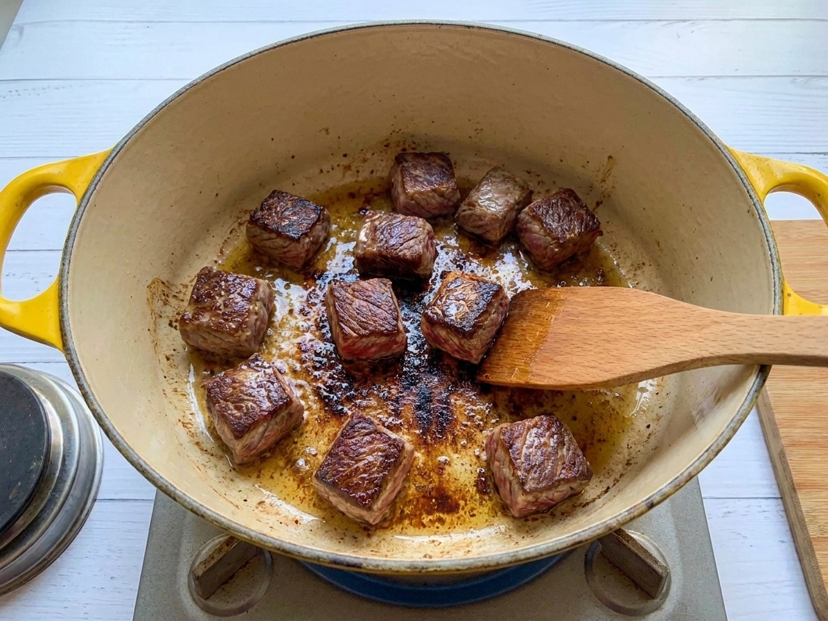 Beef cubes searing in a Dutch oven on a stovetop with browned edges and fond forming on the bottom