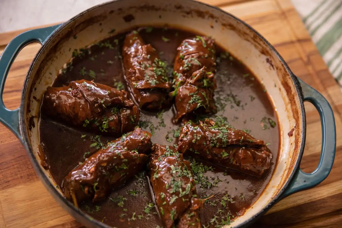 Beef rouladen rolls browning in a Dutch oven on the stovetop with tongs turning one roll and browned bits in the bottom of the pot