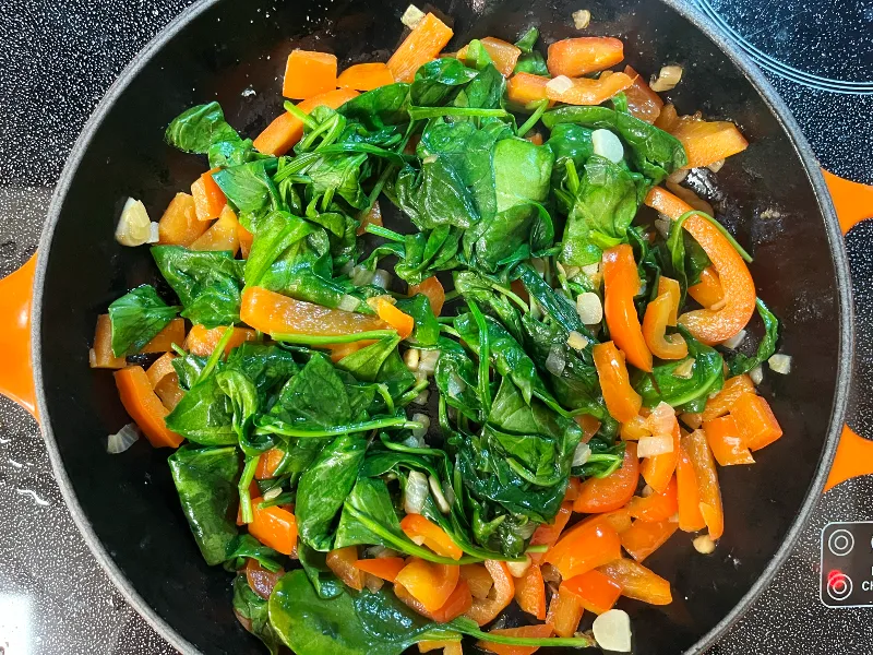 Bell peppers and spinach being sautéed in a skillet on a stovetop with a wooden spoon