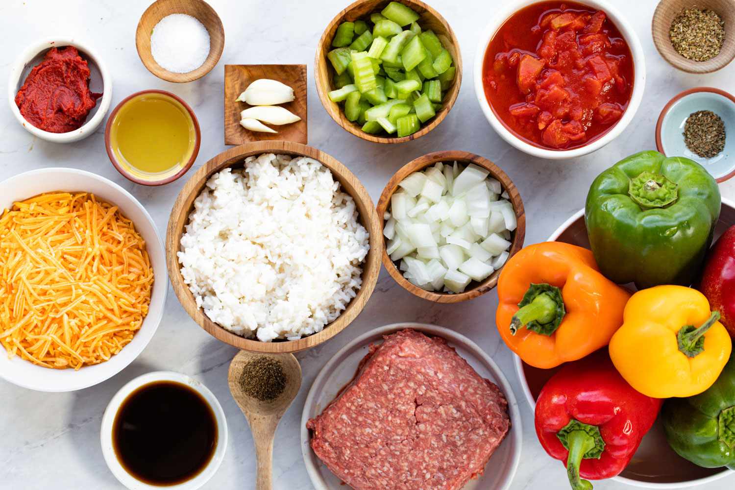 Bell peppers, cooked rice, ground beef, onion, garlic, canned tomatoes, tomato paste, shredded cheese, and spices arranged on a home kitchen counter in natural light