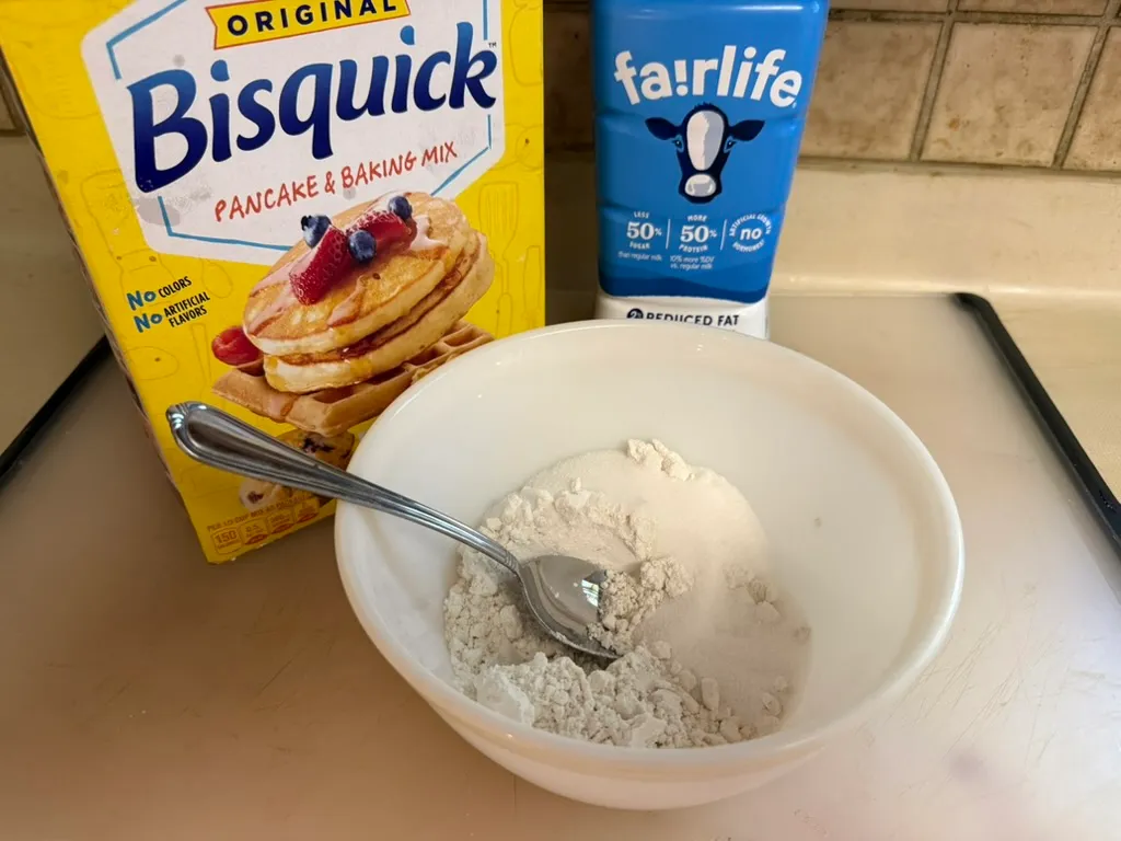 Bisquick baking mix, a measuring cup of milk, and a mixing bowl arranged on a bright kitchen counter