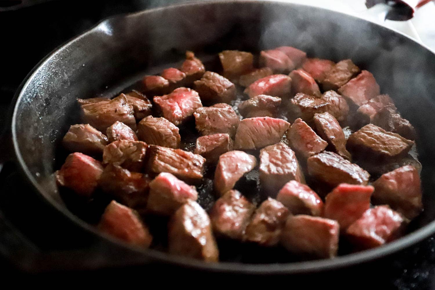 Bite-size steak pieces browning in a hot cast iron skillet with visible steam and browned edges, a wooden spoon resting nearby on the stovetop