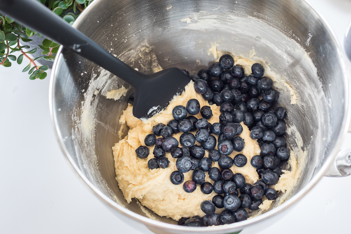 Blueberries being folded into thick lemon quick bread batter with a rubber spatula in a large mixing bowl on a home kitchen counter
