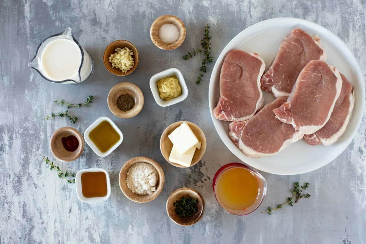 Boneless pork loin chops, kosher salt, black pepper, garlic, lemon, thyme, chicken broth, and butter arranged on a clean kitchen counter with a cutting board