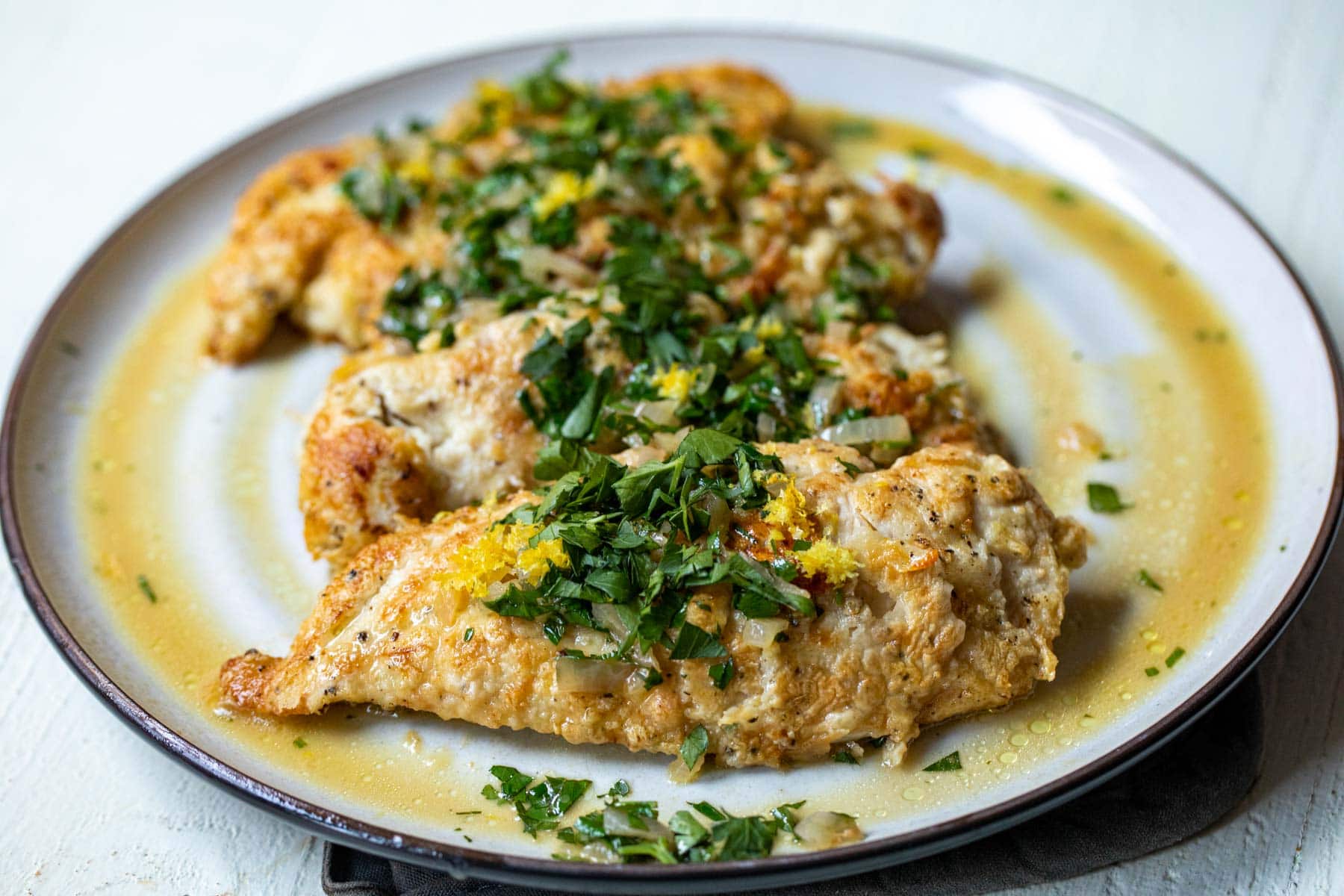 Boneless skinless chicken cutlets, lemon, garlic, butter, chicken broth, parsley, and flour arranged on a tidy kitchen counter with a small bowl of kosher salt
