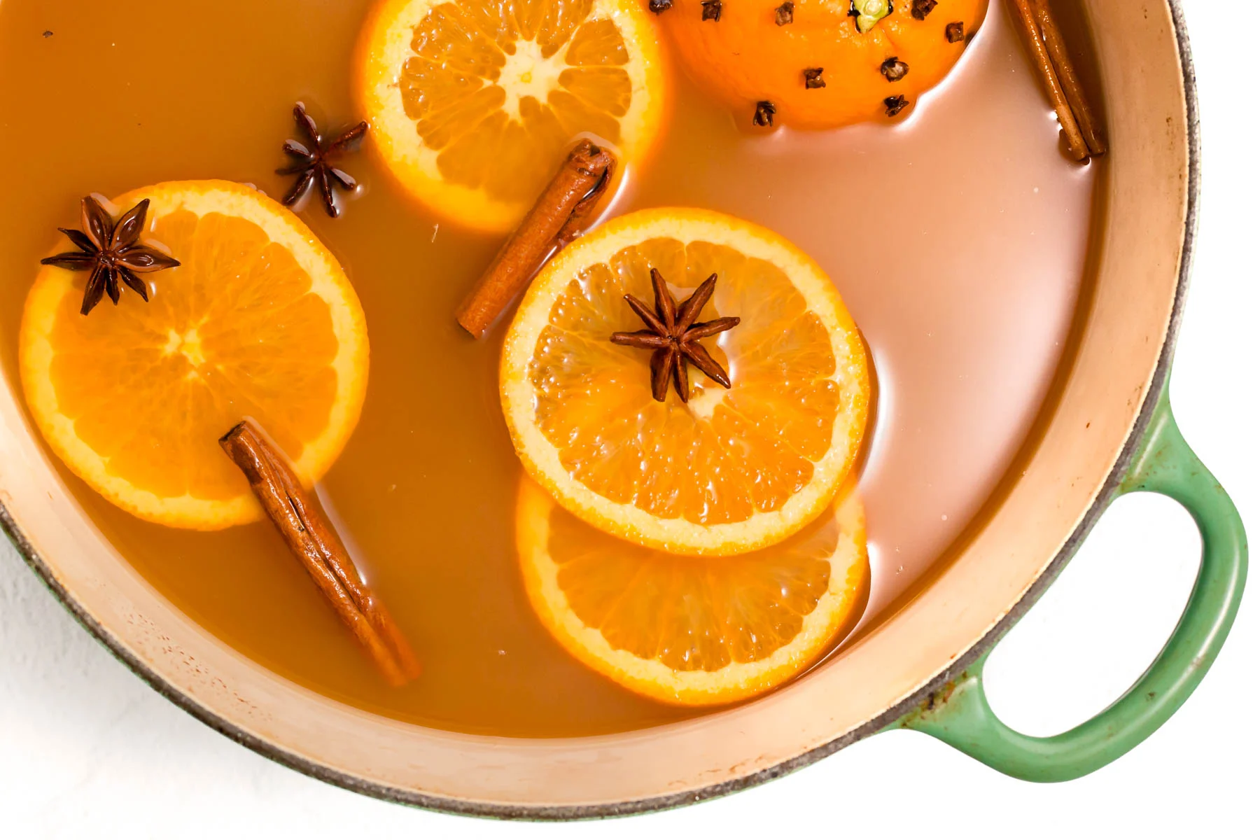 Bottles of apple cider with whole cinnamon sticks, cloves, star anise, brown sugar, and an orange arranged on a clean kitchen counter