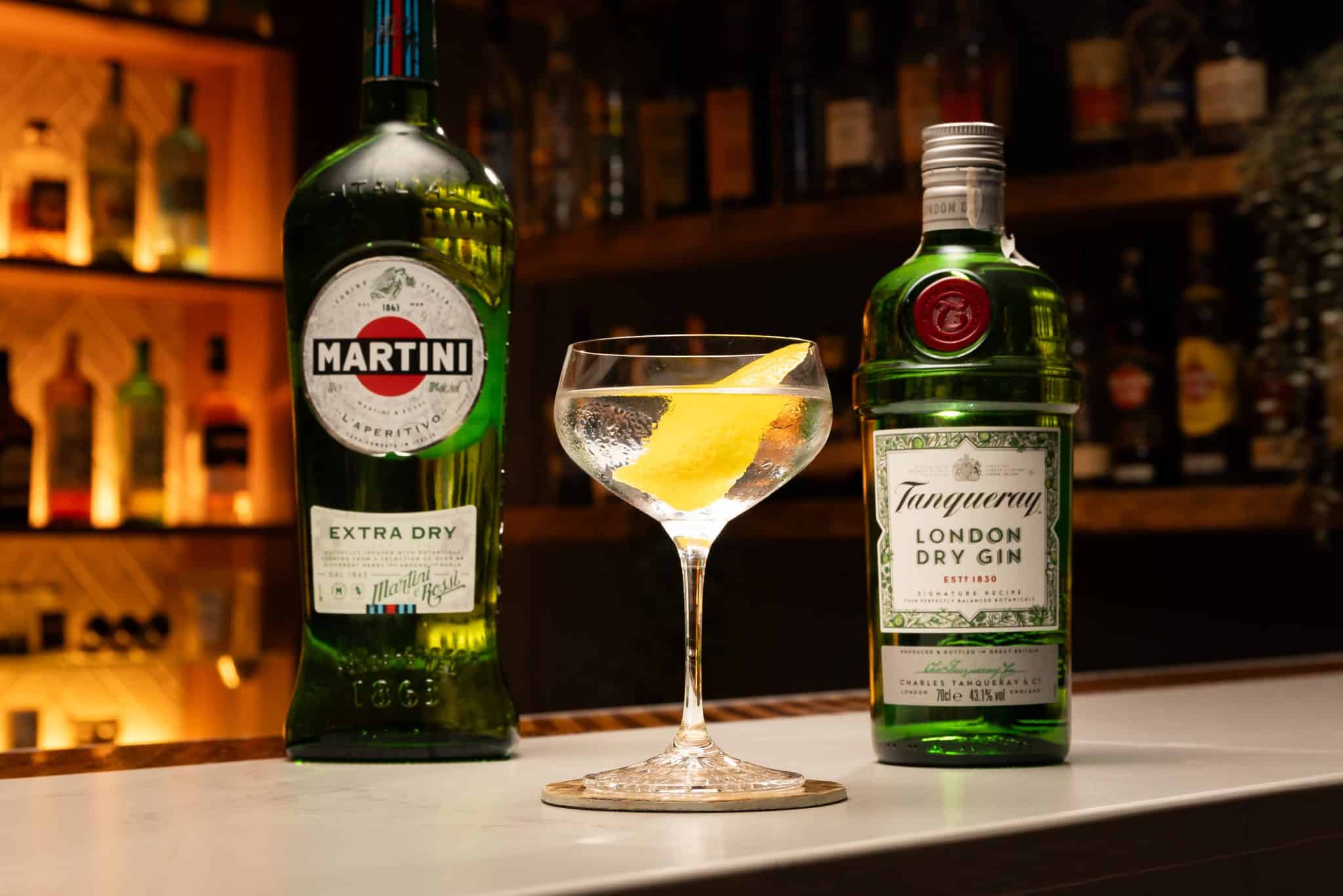 Bottles of gin and dry vermouth beside a small dish of green olives, a lemon, and a jar of cocktail onions on a tidy kitchen counter