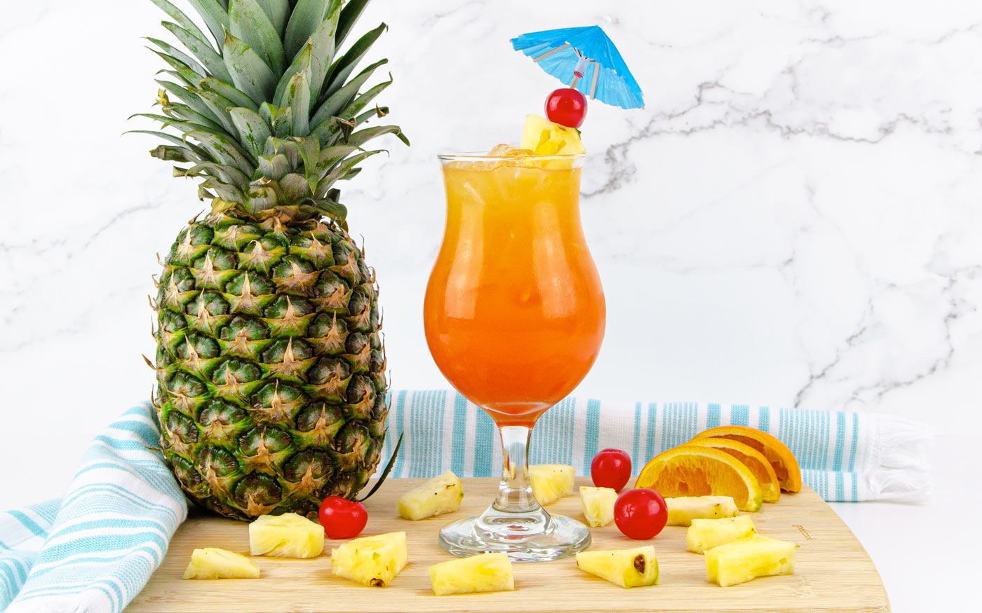 Bottles of light rum and dark rum beside pineapple juice, orange juice, lime, and a bottle of grenadine arranged on a clean kitchen counter with a cutting board and ice nearby