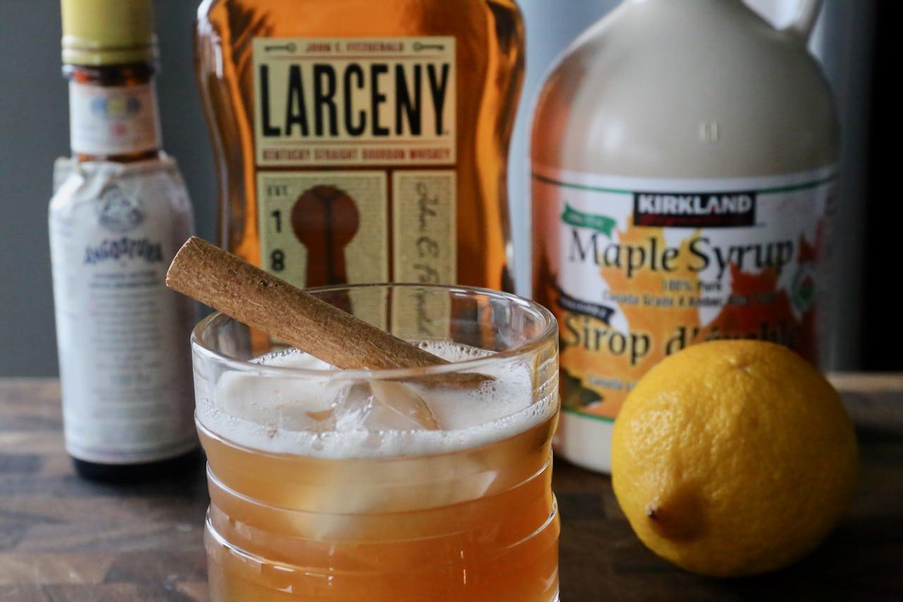Bourbon, fresh lemons, a small bottle of maple syrup, and a bottle of bitters arranged on a clean kitchen counter with a jigger and cocktail shaker
