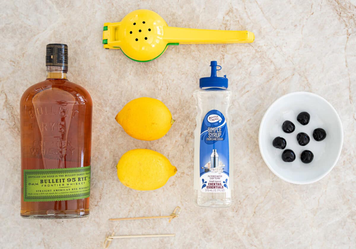 Bourbon, fresh lemons, simple syrup, Angostura bitters, and an egg arranged on a clean kitchen counter with a jigger and cocktail shaker