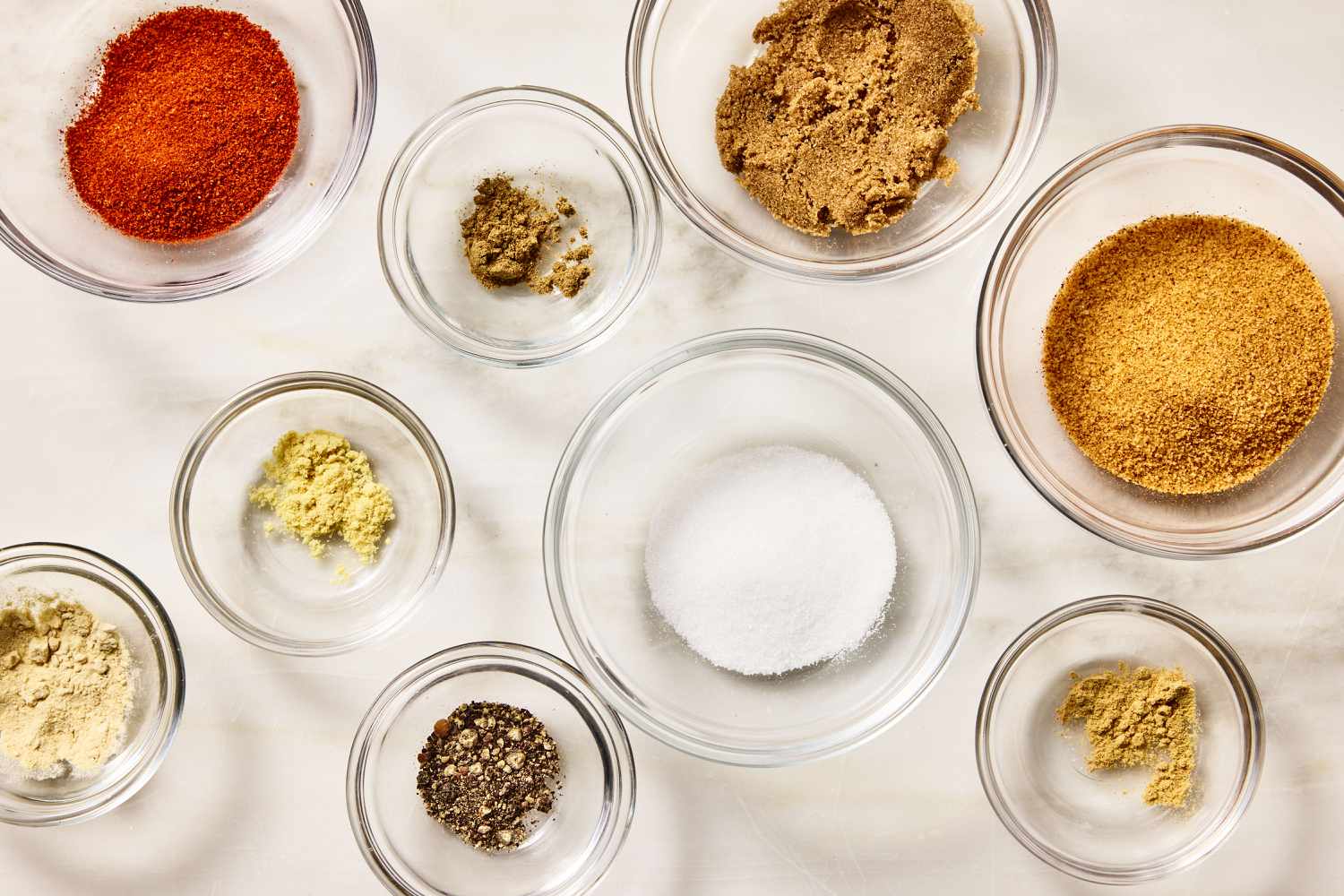 Bowls of brown sugar, paprika, salt, black pepper, garlic powder, and chili powder arranged on a kitchen counter beside a raw pork butt