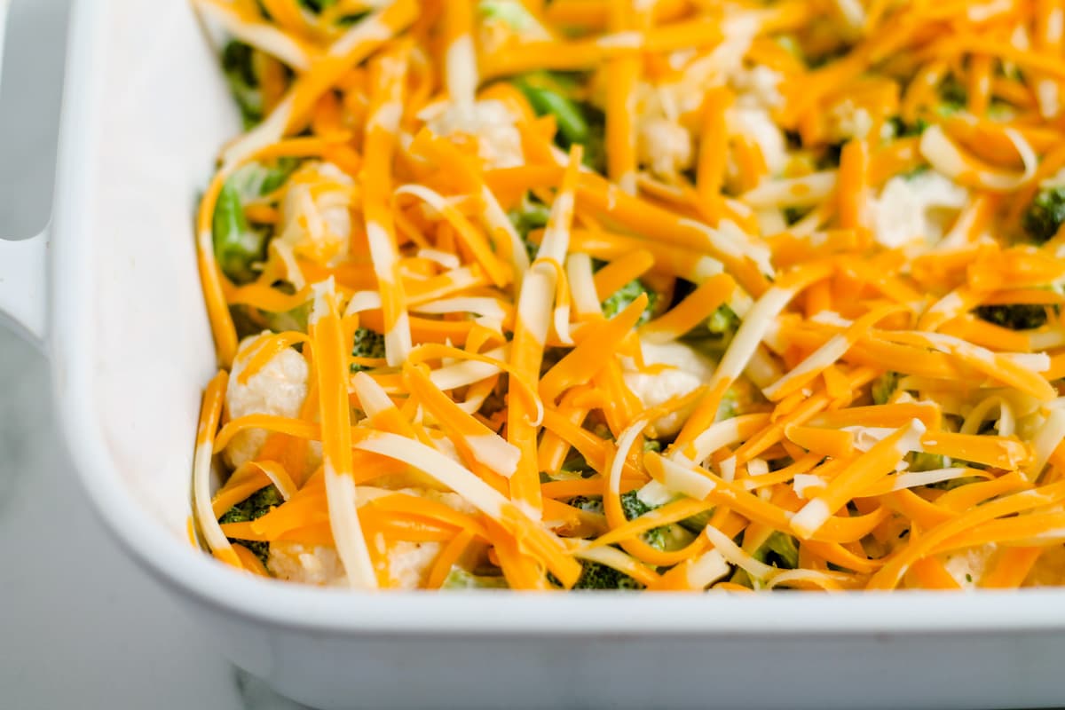 Bowls of chopped broccoli, cauliflower, carrots, and sliced mushrooms with shredded cheddar and crackers on a clean kitchen counter
