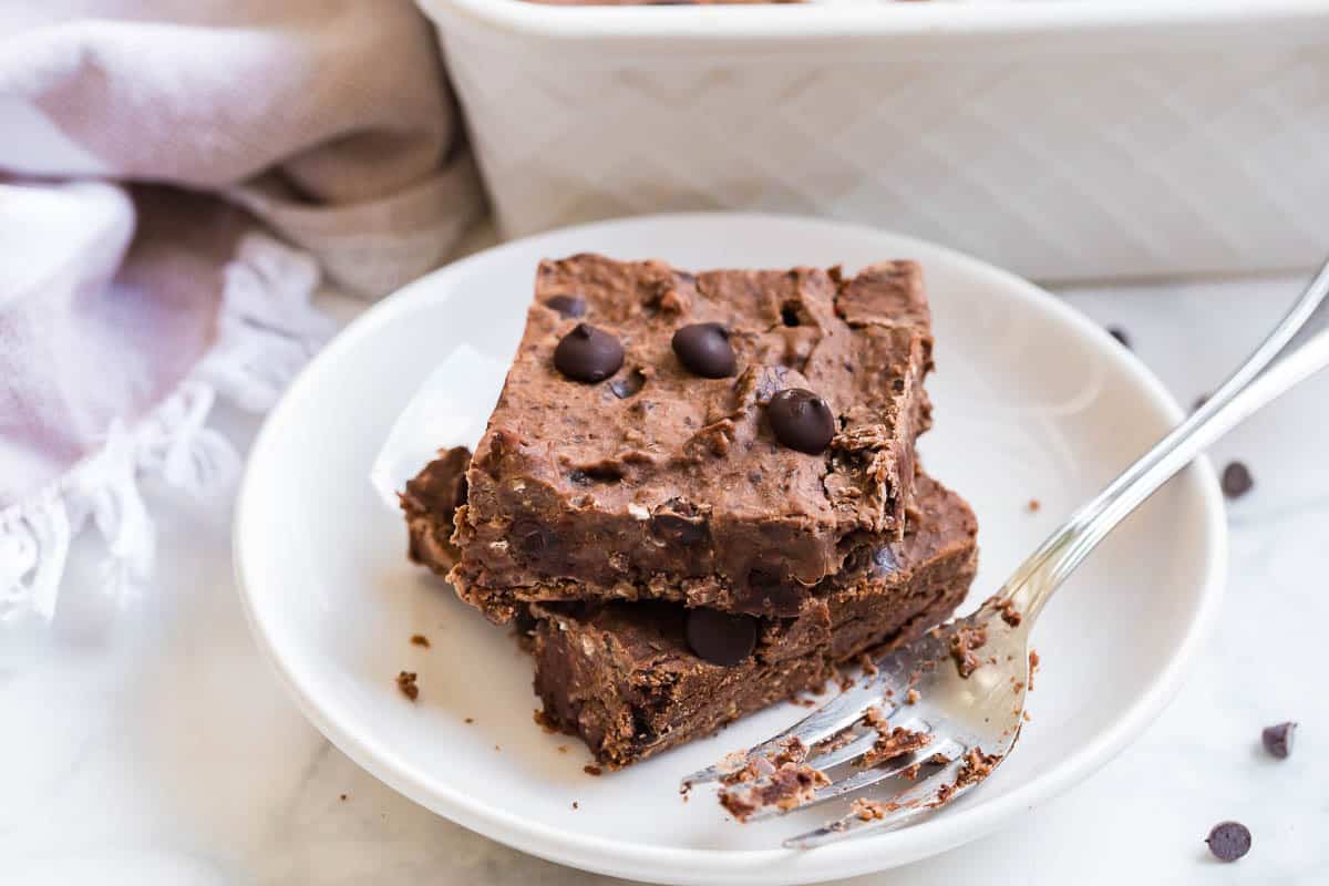 Bowls of cocoa powder, vanilla protein powder, Greek yogurt, eggs, maple syrup, chocolate chips, and baking powder arranged neatly on a kitchen counter