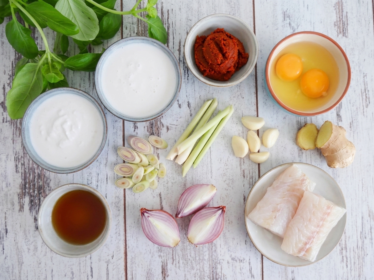Bowls of coconut milk, sliced lemongrass, garlic, shallots, ginger, curry paste, fish sauce, eggs, and white fish fillets arranged on a neutral kitchen counter