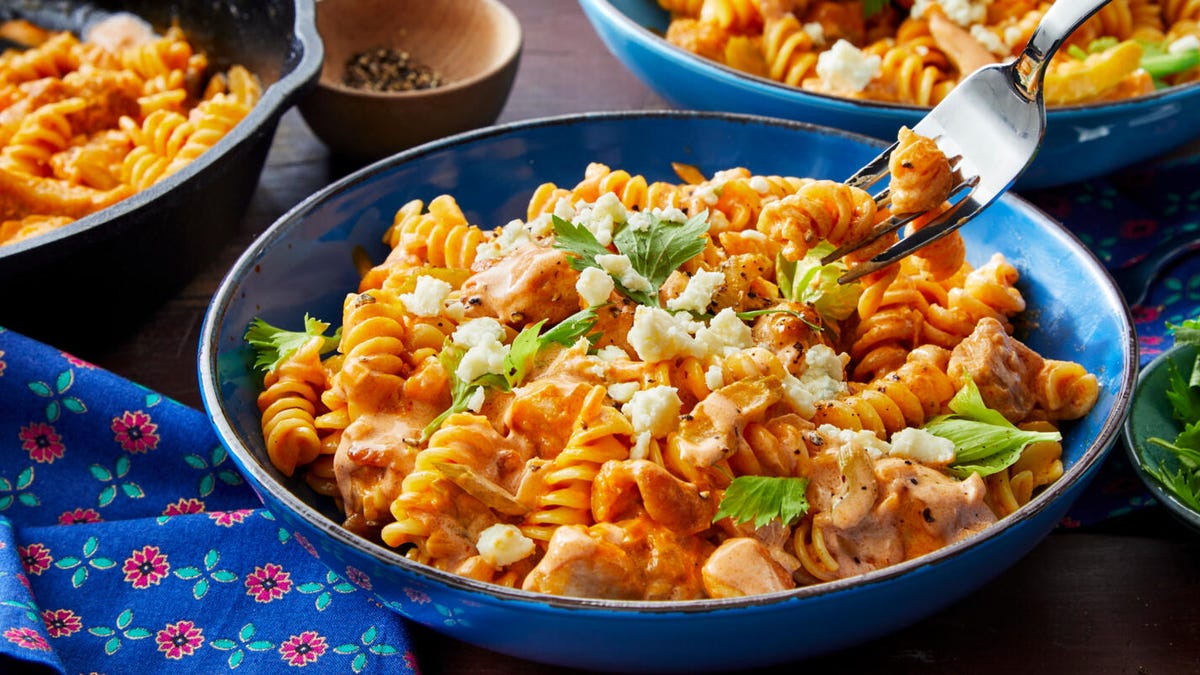 Bowls of cooked pasta, shredded chicken, Buffalo hot sauce, cream cheese, shredded cheddar, garlic, and green onions arranged on a bright kitchen counter