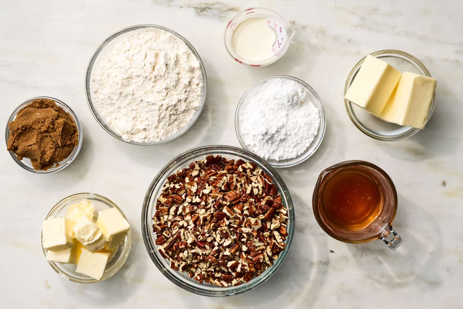 Bowls of flour, brown sugar, pecans, butter, eggs, and maple syrup arranged on a light colored kitchen counter with measuring spoons