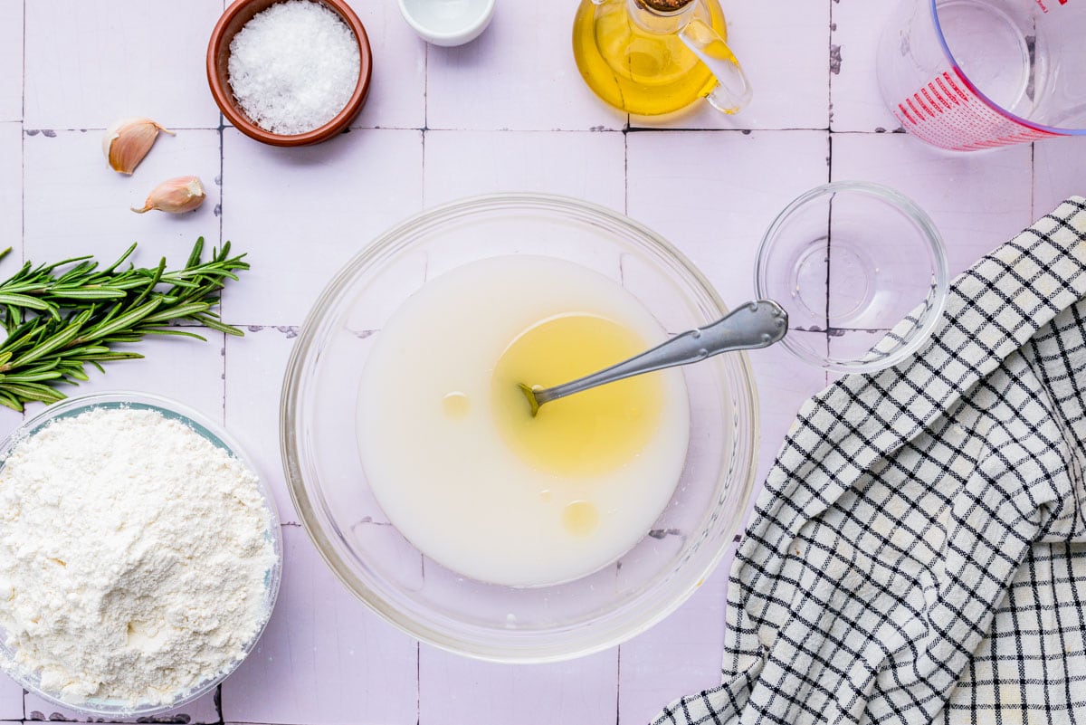 Bowls of flour, yeast, salt, olive oil, and a small bowl of chopped rosemary arranged on a bright kitchen counter