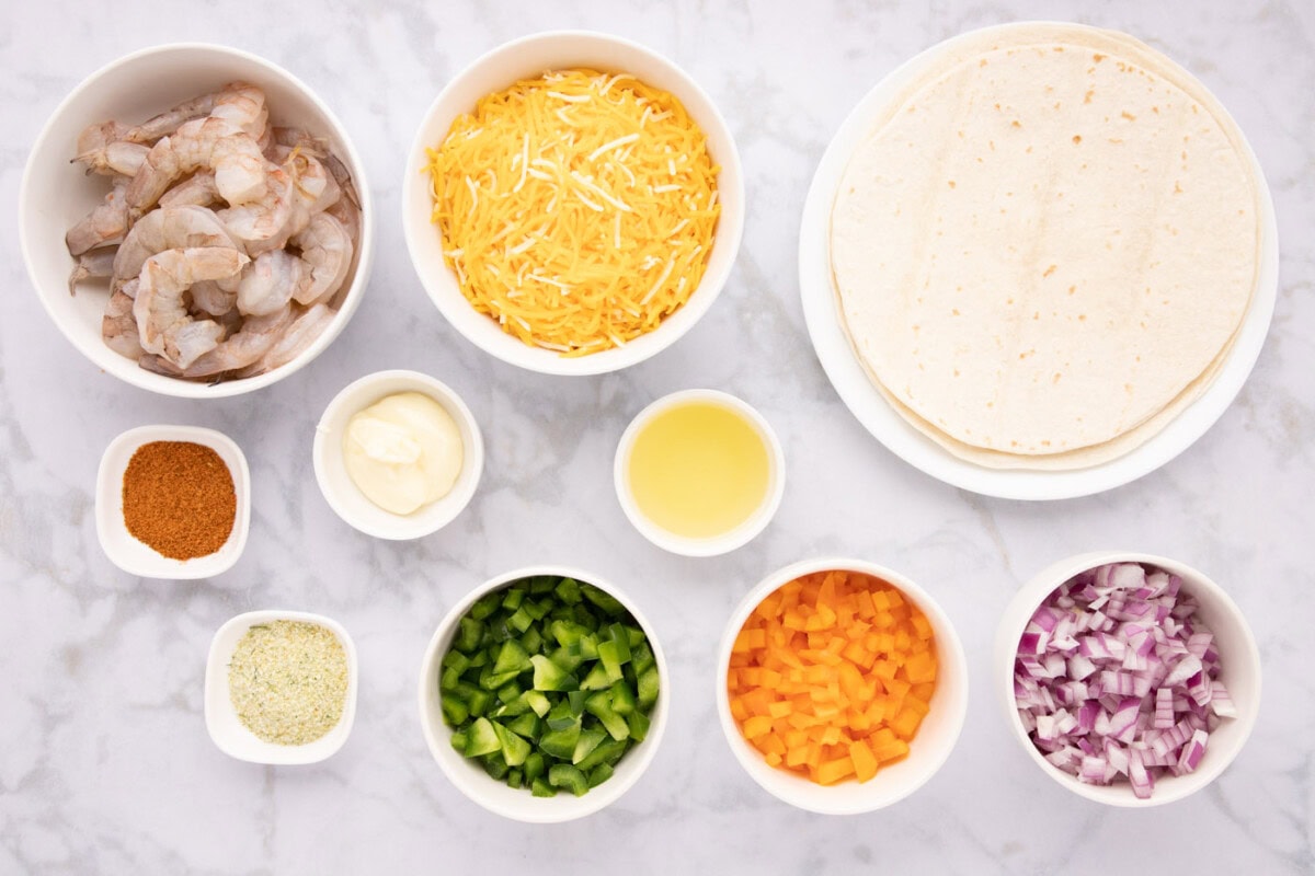 Bowls of peeled shrimp, shredded cheese, sliced peppers and onions, tortillas, lime, sour cream, and spices arranged on a tidy kitchen counter under warm light