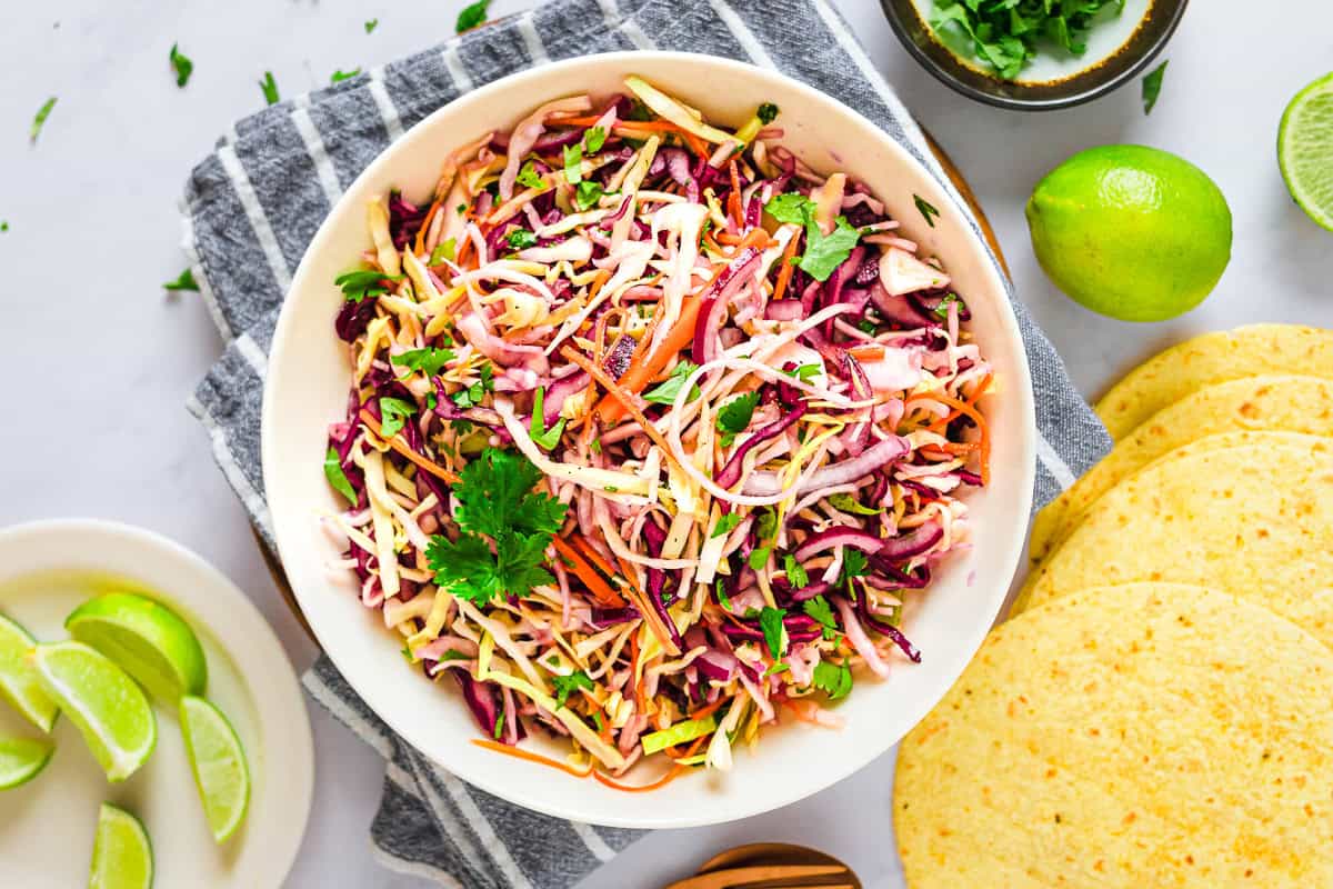 Bowls of shredded green cabbage, shredded purple cabbage, grated carrots, chopped cilantro, sliced jalapeno, and limes arranged on a bright kitchen counter