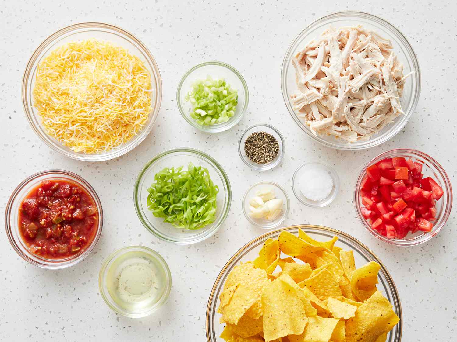 Bowls of tortilla chips, shredded cheddar, shredded Monterey Jack, cooked taco meat, black beans, diced tomato, sliced jalapeños, and chopped cilantro arranged on a kitchen counter