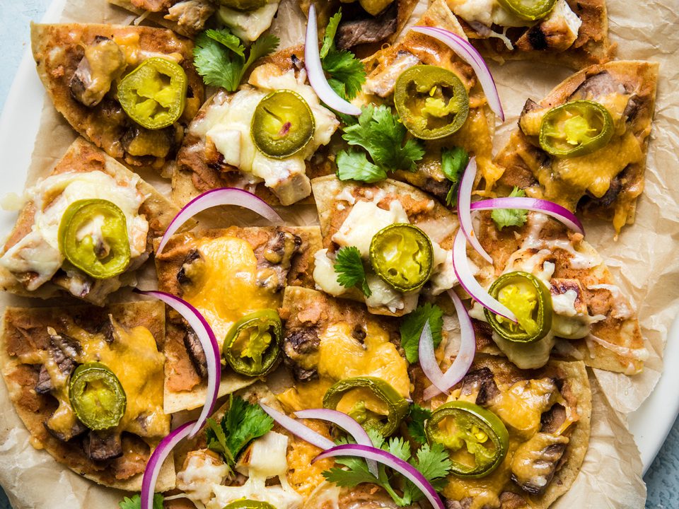 Bowls of tortilla chips, shredded cheese, cooked ground beef, sliced jalapeños, diced tomatoes, and chopped cilantro arranged on a bright kitchen counter